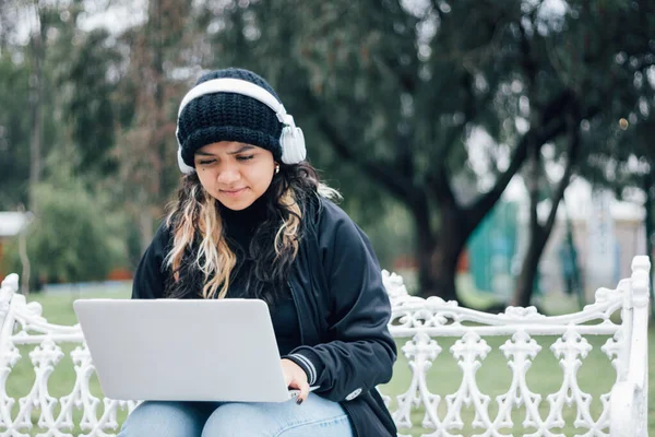 Latin üniversiteli bir kız parkta bir bankta dizüstü bilgisayar kullanarak oturuyor.