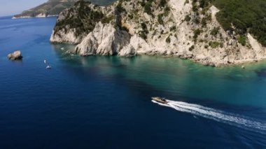 Kayalık yeşil dağ boyunca ilerleyen lüks bir tekne, turkuaz su. Arkadan ve yukarıdan yarım daire drone uçuşu. Kayayla küçük bir kaya arasına girer..