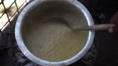 Black hand stirs liquid whitish porridge in an iron bowl with iron scoop..