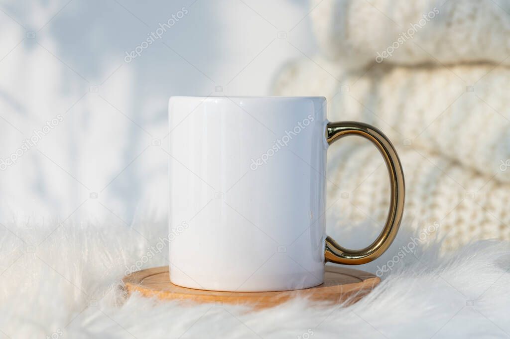 White ceramic coffee mug mockup with cozy winter background with warm sweaters and leather rug and copy space for your design. Blank sapce for promotional content. Standart mug with golden handle