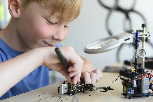 Niño inventor sonriente joven que ensambla cuidadosamente el juguete robótico con el destornillador, trabaja con pequeñas piezas mecánicas, usando lupa. Educación STEM Imágenes de stock libres de derechos