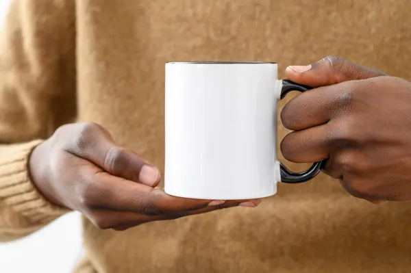 Taza de cerámica maqueta. Cierre las manos afroamericanas sosteniendo taza blanca con mango negro de café, espacio para copiar. Vista frontal, espacio para impresión de marca Fotos de stock