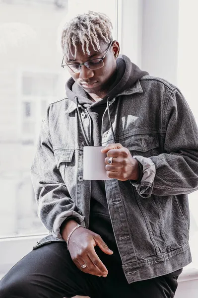 Um modelo de caneca de cerâmica. Retrato de afro-americano em uma jaqueta de ganga segurando copo branco com alça preta de café, espaço de cópia. Vista frontal, espaço para a impressão da marca — Fotografia de Stock