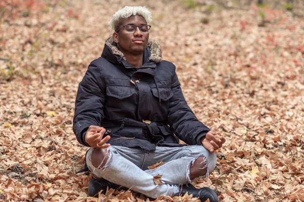 Armonía consciente y concepto de relajación. Joven hombre negro respirando aire fresco con los ojos cerrados meditando en el parque de otoño al aire libre Fotos de stock