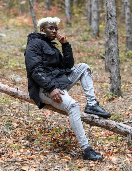La naturaleza otoñal con la humana. Joven afroamericano descansando en el bosque, sentado en un árbol caído — Foto de Stock