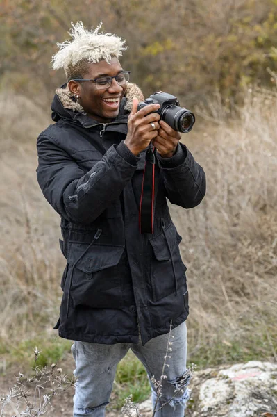 Retrato de comprimento total do jovem afro-americano bonitão hipster caminha em um parque de outono no fundo de folhas amarelas e tira fotos em uma câmera profissional — Fotografia de Stock