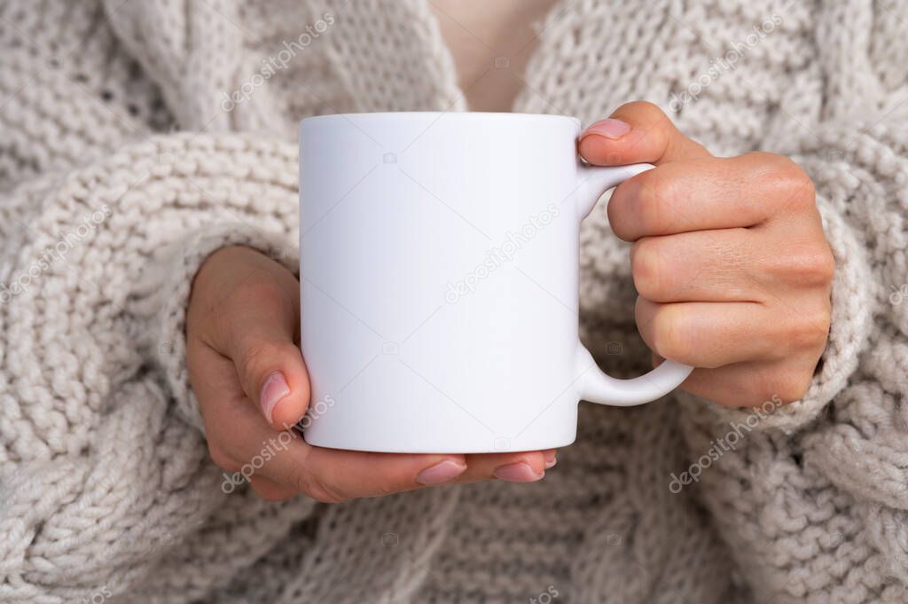 Woman in knitted sweater holding white mug in hands. Winter cup Mockup for Christmas individual gifts design