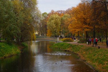 Park yerinde sıraya girin. Sonbahar doğasının güzel renkleri. Yeşil, sarı ve kırmızı ağaçlar. Nehrin üzerindeki kemerli köprü. İnsanlar arka planda parkta yürürler.