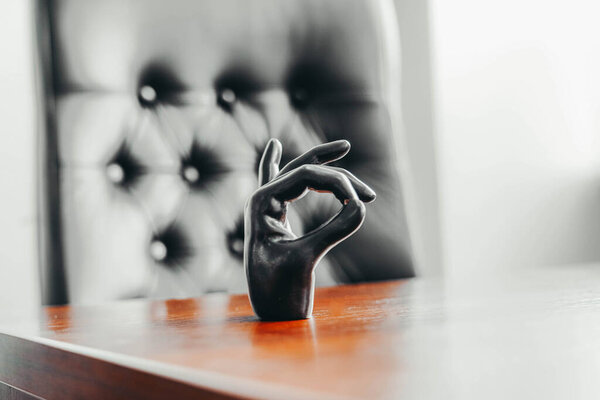 Photo of a black palm hand showing an okay sign stands on a wooden brown table near a black leather chair in the office.