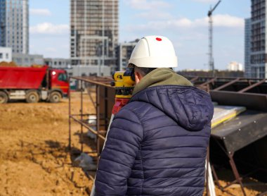 Beyaz miğferli bir inşaat işçisinin fotoğrafı. Şehrin caddesinde kırmızı kamyonlu, çok katlı binaların inşaat alanına bakıyor..