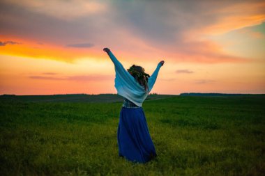 Tarlada, yeşil çimenli ve günbatımlı güzel bir tarlanın arka planında, uzun sarı saçlı, şık mavi etekli bir çiçek çelengi içinde genç bir kadının fotoğrafı.