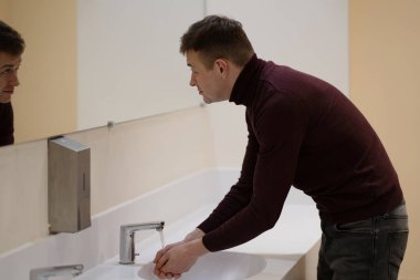 Umumi tuvalette ellerini yıkayan ve aynada kendine bakan genç bir adamın fotoğrafı.