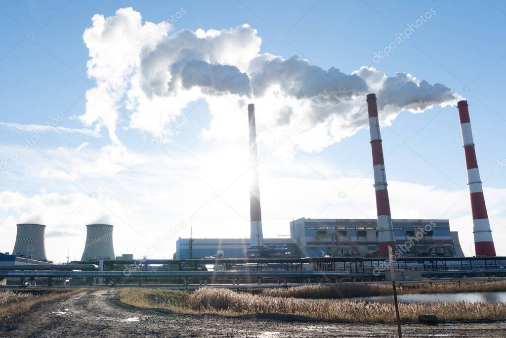 Photo of three chimneys from which thick white smoke comes out against a background of blue sky and sun