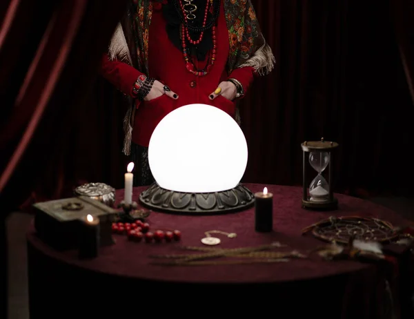 Astroloji için objelerle dolu bir masa. Mumları yakan kum saati ve arka planda parlak giysiler içinde kadın astrolog ve cepleri dolu eller.