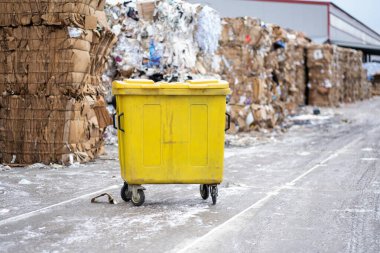 Bahçeye sıkıştırılmış kağıt balya yığını. Arka planda çöp ve kağıt dolu sarı bir çöp konteynırının fotoğrafı.