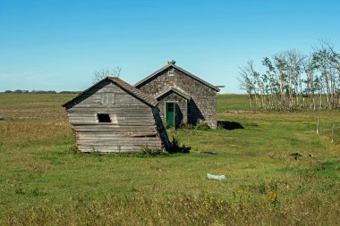 Yıpranmış, terk edilmiş bir ev ve Kanada, Saskatchewan kırsalında eski bir çiftlik bahçesinde eğilmiş bir kulübe..