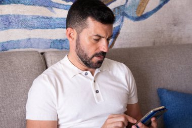 Otel odasındaki kanepede oturan adam cep telefonuyla iletişim kuruyor..