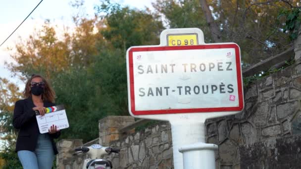 Žena s filmovým clapperboardem kráčející směrem k ceduli Saint Tropez. — Stock video