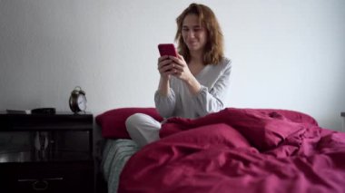  Genç bir kadın pijamalarıyla yatakta oturuyor ve cep telefonuyla iletişim kuruyor..
