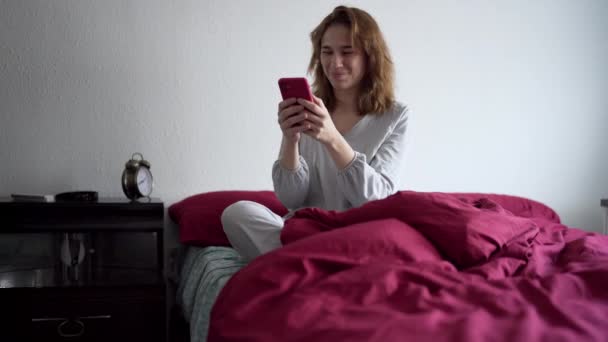 Ung kvinde sidder i sengen i pyjamas interagerer med mobiltelefon. – Stock-video