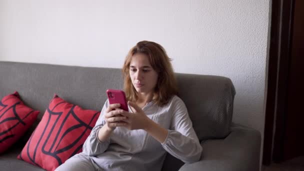 Jovem mulher sentada no sofá de pijama interagindo com o telefone móvel. — Vídeo de Stock