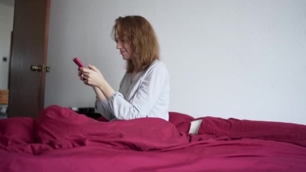 Jonge vrouw zitten in bed in pyjama interactie met mobiele telefoon. — Stockvideo