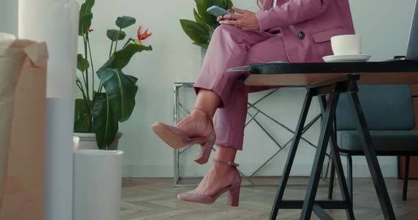 Ontspannen op het werk. Close-up benen van elegante zakenvrouw in roze pak zitten op kantoor tafel met behulp van mobiele telefoon app. — Stockvideo