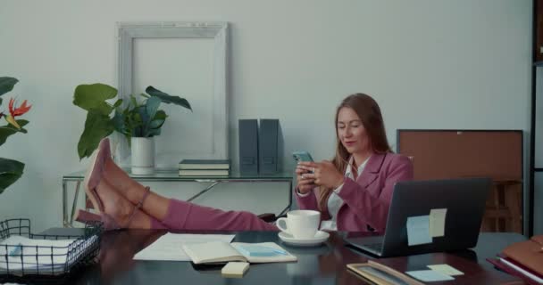 快適なショッピングオンライン。オフィスのテーブルの上に足で携帯電話アプリを使用して美しい幸せな女性の上司. — ストック動画