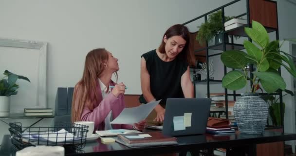 Trabajando juntos. Dos hermosas mujeres de negocios jóvenes de 30 años hablan con el ordenador portátil que coopera en la mesa de oficina moderna luz. — Vídeo de stock