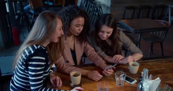 Concepto de conexión móvil. Tres jóvenes felices sonrientes amigas multiétnicas usan las redes sociales de teléfonos inteligentes en la cafetería. — Vídeos de Stock