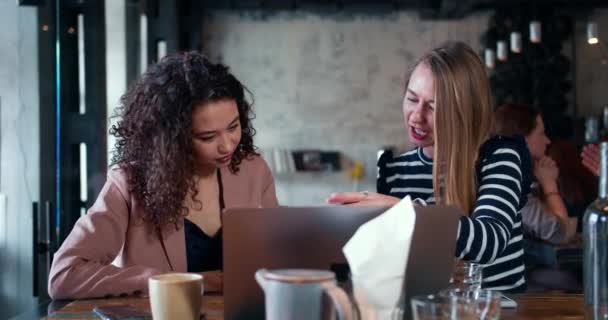 Teamwork concept. Twee gelukkige aantrekkelijke multi-etnische zakenvrouwen doen papierwerk praten achter laptop bij coffeeshop. — Stockvideo