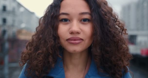 Close-up portrait of beautiful happy mixed race woman smiling at camera, touching curly hair at city bridge slow motion. — Stock Video