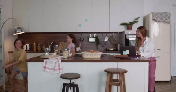 Noisy děti pobíhají kolem svobodná maminka obchod žena se snaží soustředit a pracovat na prostorné domácí kuchyně stůl pomocí notebooku. — Stock video