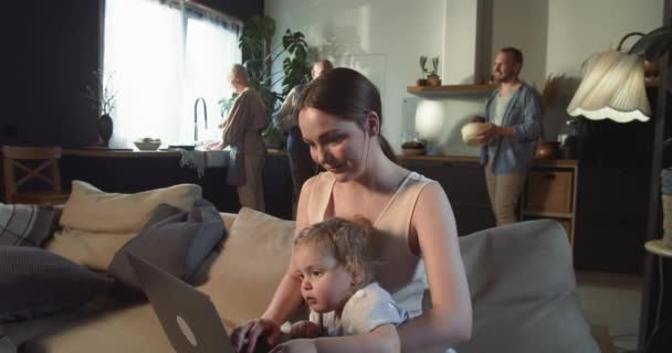 Vergrößern Sie glückliche heitere Mutter mit Laptop-Shopping online mit Baby-Mädchen, Kindern und älteren Verwandten im Hintergrund zu Hause. — Stockvideo