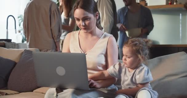 Glad vacker ung 20-tals mamma använder laptop tillsammans med söt liten flicka sitter på soffa med stor familj i vardagsrummet. — Stockvideo