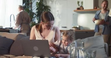 Mutlu gülümseyen 20 'li yaşlardaki genç anne internette tatlı bir kızla alışveriş yapıyor dizüstü bilgisayarını kullanıyor ailesiyle oturma odasında oturuyor.