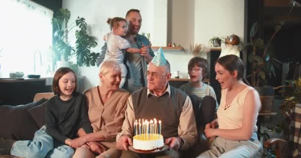 Happy old senior grandfather celebrating birthday with cake and confetti together with wife, kids and grandkids at home. — Stock Video