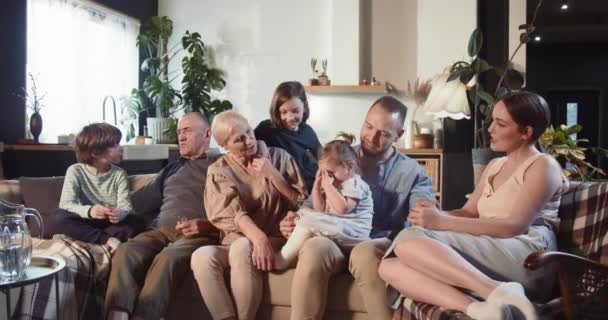 Boldog vidám idős nagyszülők, fiatal apa és anya, tini gyermekek és a kislány kötődés kanapén a fény otthon. — Stock videók
