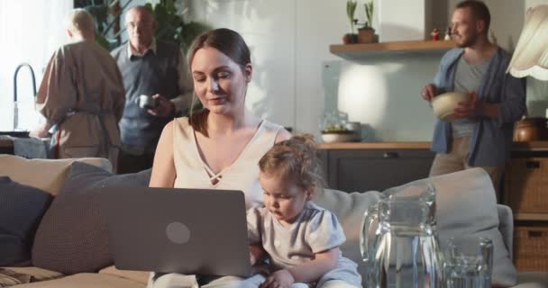 Glücklich attraktive junge Mutter mit Laptop zusammen mit niedlichen Baby-Mädchen Online-Shopping zu Hause, Familie im Hintergrund. — Stockvideo