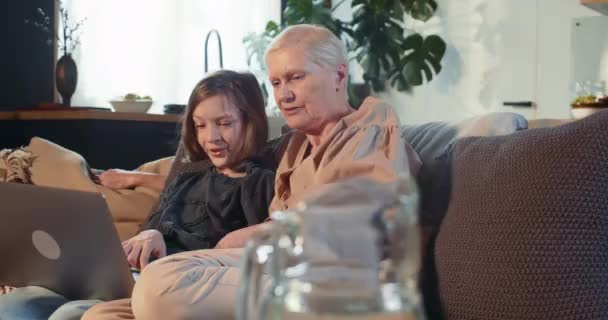 Щаслива усміхнена дівчина-підліток навчає старшу бабусю онлайн безпеці, використовуючи ноутбук, що сидить на домашньому дивані. Догляд за літніми людьми . — стокове відео