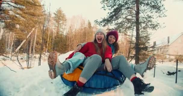 Два мультиэтнических взволнованных счастливых красивых друзей женщины улыбаются сани на склоне снега к камере, зимнее удовольствие замедленной съемки. — стоковое видео