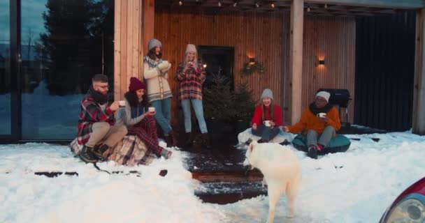 Grupa szczęśliwych, zrelaksowanych, wieloetnicznych przyjaciół pije gorącą herbatę w przytulnym zimowym domu taras z psem, czerwony samochód zwolniony ruch — Wideo stockowe