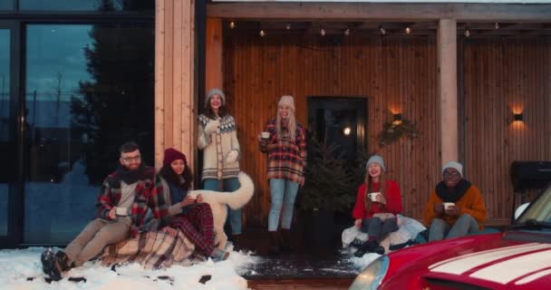 Schöne Weihnachtsferien. Gruppe fröhlicher Freunde mit Heißgetränken blickt auf der Winterhausterrasse in Zeitlupe in die Kamera. — Stockvideo