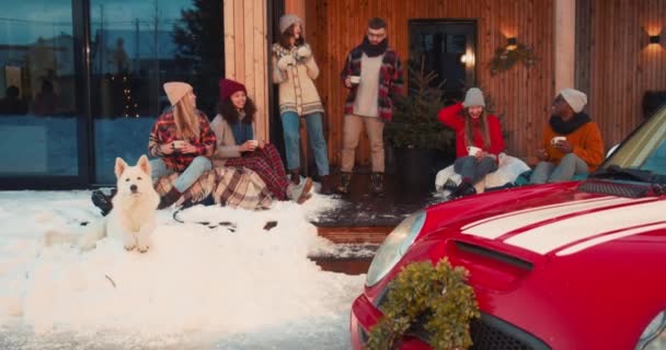 Fête de Noël en plein air. Ami joyeux et heureux dansent au chalet d'hiver enneigé confortable maison avec chien, voiture rouge au ralenti. — Video