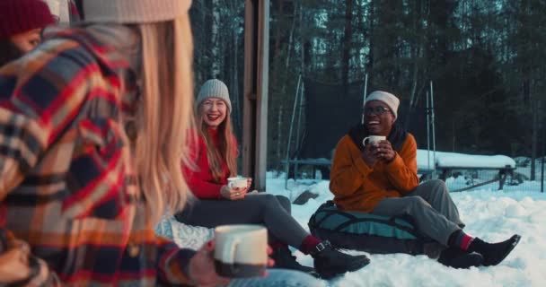 Веселые счастливые многонациональные друзья пьют чай, разговаривают улыбаясь сидя на бобовых мешках на уютной террасе зимнего дома замедленное движение. — стоковое видео