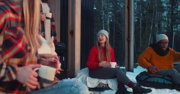 Festa de Natal. Amigos positivos felizes com bebidas quentes conversam sentados em sacos de feijão no acolhedor terraço de inverno câmera lenta. — Vídeo de Stock