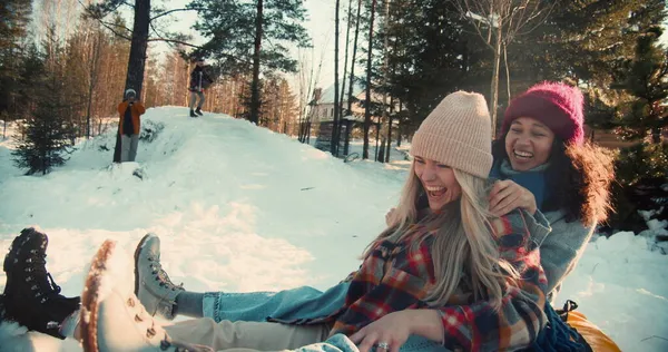 Dos atractivas mujeres felices multiétnicas sonríen en trineo en la pendiente de la nieve hacia la cámara, diversión de fin de semana de invierno cámara lenta. Imagen De Stock