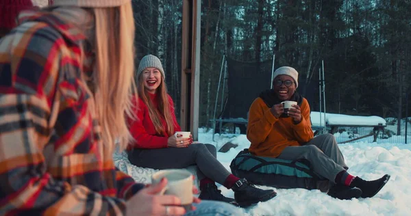 Cheerful happy multiethnic friends drink tea, talk smiling sitting on beanbags at cozy winter house terrace slow motion. — Stock Photo, Image