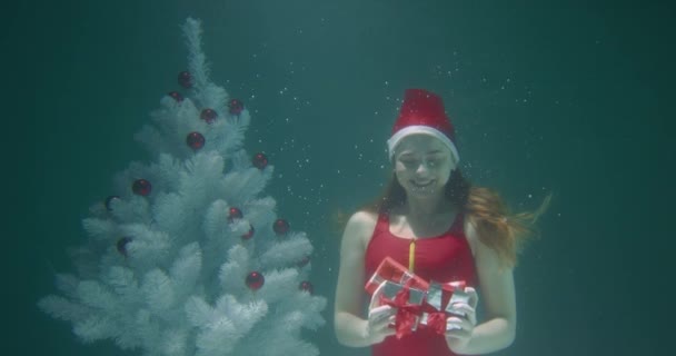 PREZENTY CHRISTMAS Szczęśliwa zabawa uśmiechnięta młoda kobieta w czerwonym stroju kąpielowym otwiera dłonie prezentami pod wodą zwolnione tempo. — Wideo stockowe