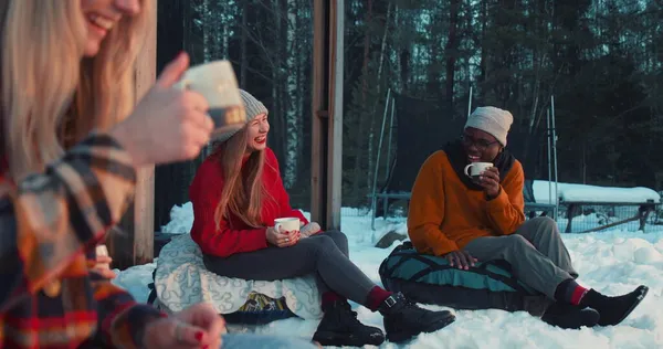 Cheerful happy multiethnic friends drink tea, talk smiling sitting on beanbags at cozy winter house terrace slow motion. — Stock Photo, Image